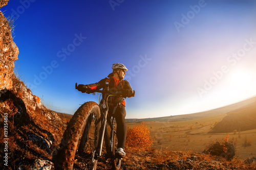 Cyclist man standing on a hill with bicycle and enjoying landscape on a sunny day against a blue sky