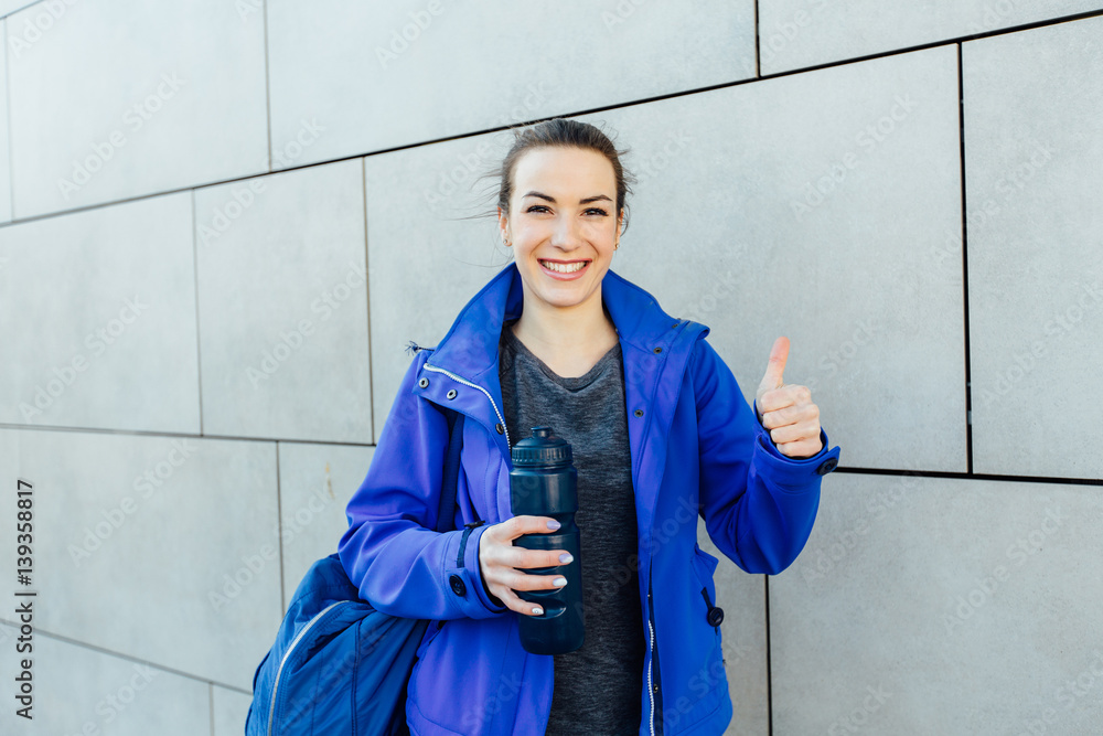 Joyful attractive sporty woman in blue sportswear holding bottle and show thumb up in front of the wall. Joyful attractive sporty woman in blue sportswear holding bottle and show thumb up in front of the wall.