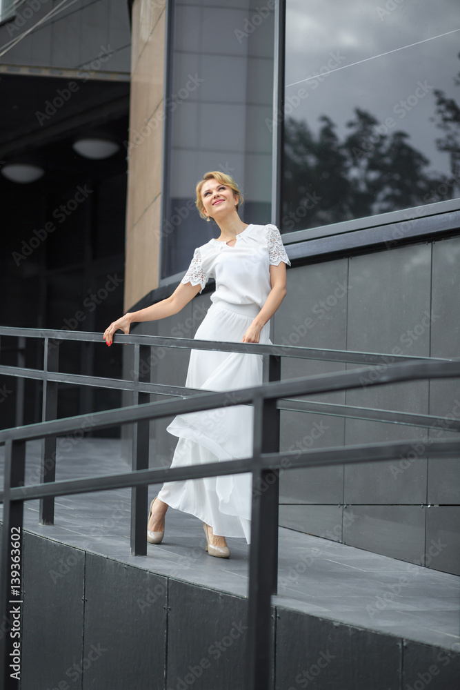 Blond hair girl in white dress stands on the metal stairs, on the gray background wall. Smiling and posing in front of a window. Blond hair girl in white dress stands on the metal stairs, on the gray background wall. Smiling and posing in front of a window.