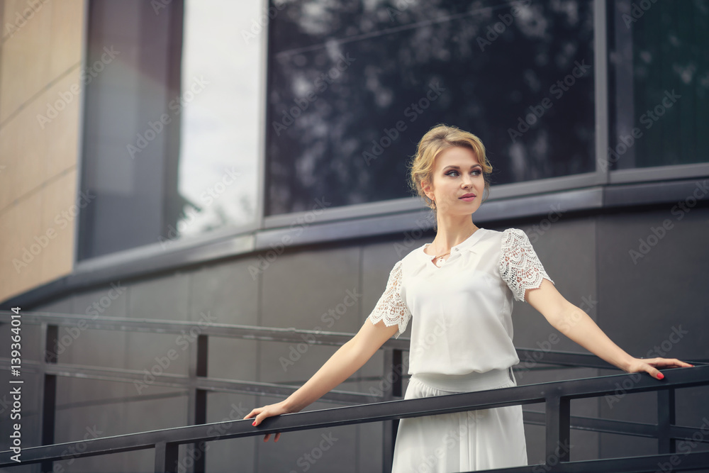 Blond hair girl in white dress stands on the metal stairs, on the gray background wall. Smiling and posing in front of a window.