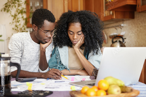 Upset young African-American couple feeling unhappy because they can't afford buying new car, facing financial problem and economic crisis, trying to solve troubles, reviewing their finances at home