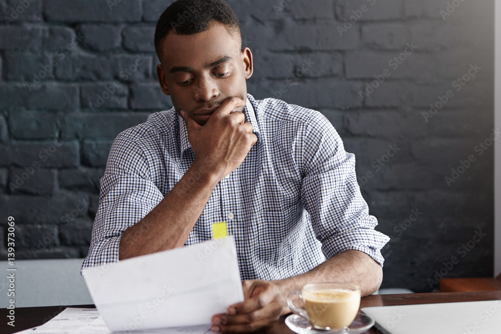 Indoor portrait of handsome serious dark-skinned businessman in formal checkered shirt working trough papers, studying document in his hands with concentrated expression, holding hand on chin Indoor portrait of handsome serious dark-skinned businessman in formal checkered shirt working trough papers, studying document in his hands with concentrated expression, holding hand on chin