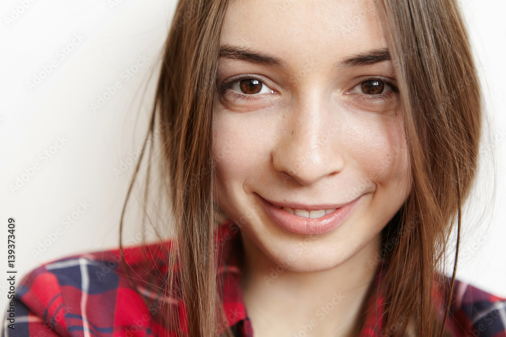 Naklejka premium Highly-detailed close up portrait of beautiful Caucasian teenage girl with dark hair, porcelain skin and brown eyes looking at camera with charming happy smile, dressed in trendy checkered shirt