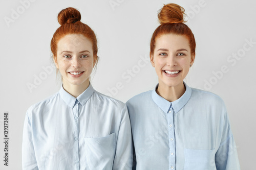 Two young redhead Caucasian females looking alike wearing same formal light-blue shirts, looking at camera, smiling happily, standing close to each other against white studio wall background photo