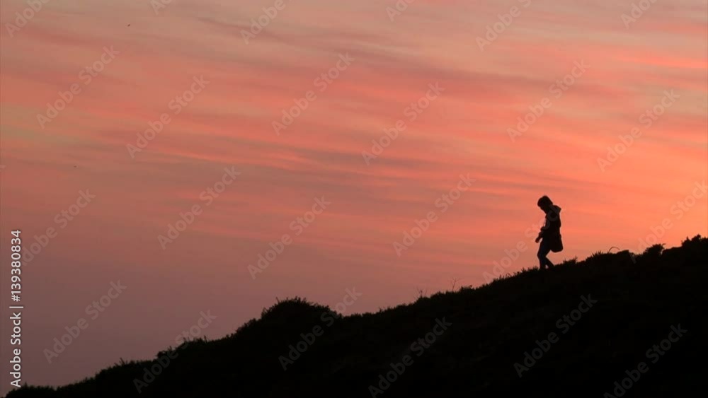 Photographer man silhouette taking pictures and going down by mountain hill, black silhouette on orange sunset sky background