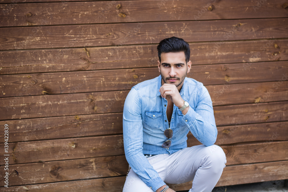 Attractive young man sitting against wood wall, looking at camera Attractive young man sitting against wood wall, looking at camera