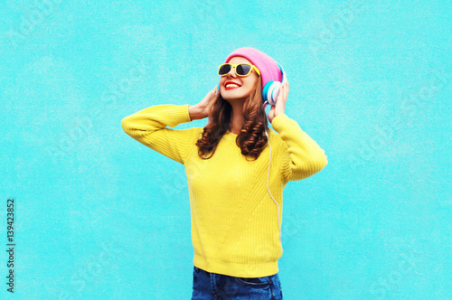 Fashion girl listening to music in headphones wearing a colorful pink hat, yellow sunglasses and sweater over blue background