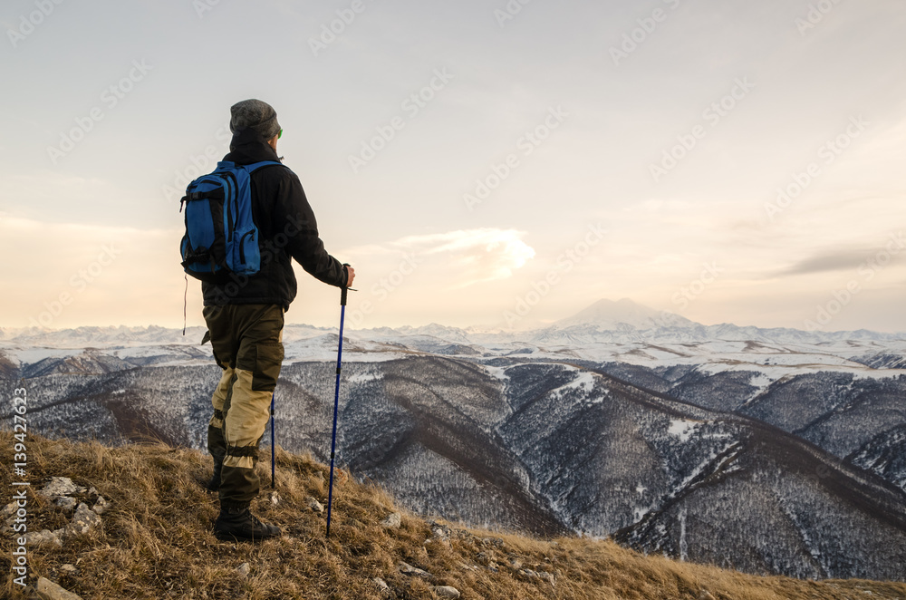 Young bearded hipster wearing a hat and sunglasses with Nordic walking sticks and a backpack moves in the mountains at sunset, against a background of sunset sky and volcano Elbrus
