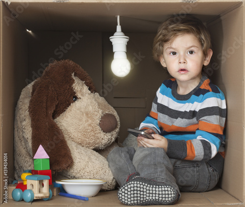Cute Toddler Boy with his Stuffed Toy in Cardboard Box with Bare Light Bulb, representing cramped small place