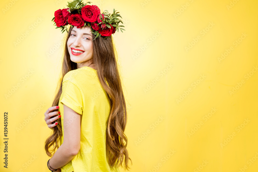 Fototapeta premium Colorful portrait of a beautiful woman in yellow t-shirt with wreath made of red roses on the yellow background