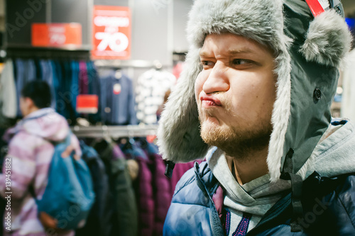 Portrait of a man with a beard with a mustache, putting on winter spring knitted hats in the store, with an emotional face shopping