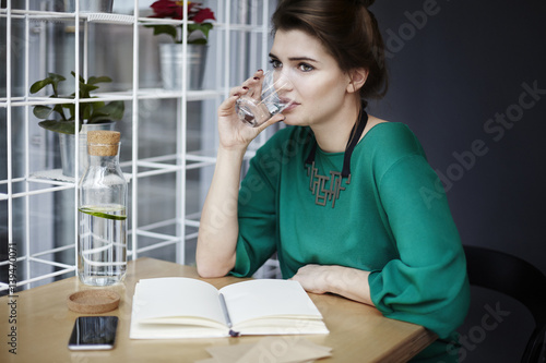 Beautiful young woman wearing green drinking pure water in cafe, having breakfast, opened book spread on table