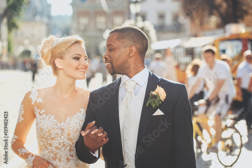 Fényképezés Happy african American groom and cute bride walking on street