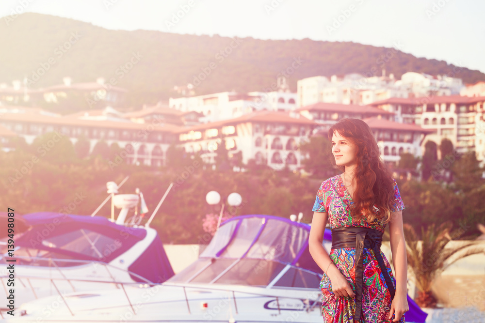 attractive brunette in long colorful dress standing alone on the beach against the backdrop of yachts and town at sunset