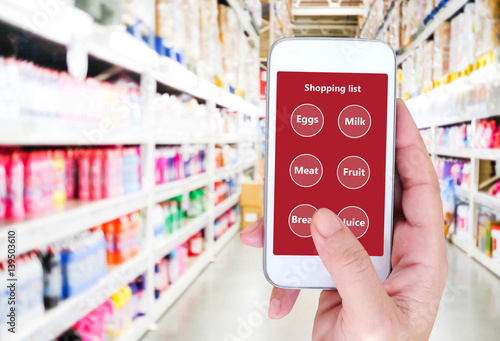 Hand holding smart phone with grocery shopping check list application on screen over blur product shelves in supermarket background, business and technology concept