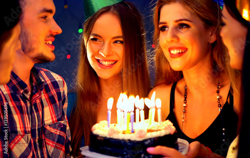 Happy friends birthday celebrating food with candle celebration cakes in club. People wear in hat party looking at burning candles. Women and men have fun.