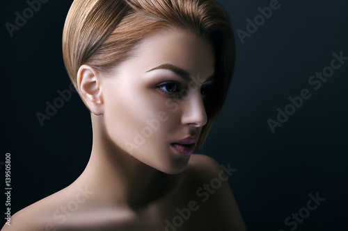 Indoor close up portrait of a young beautiful woman with healthy short hair posing on dark background, part of face in shadow. Model looking forward, aside. Female beauty and advertisement concept. 