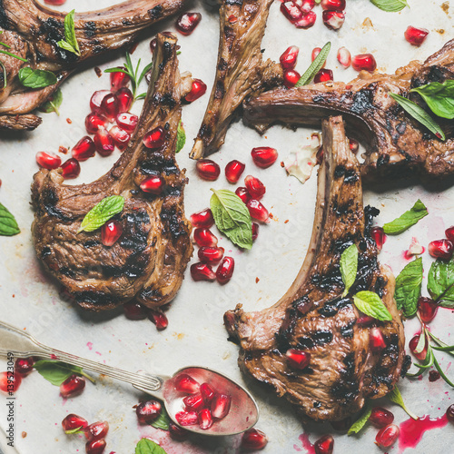 Fototapeta Naklejka Na Ścianę i Meble -  Grilled lamb ribs with pomegranate seeds, fresh mint and rosemary over metal baking tray background, top view, square crop. Meat barbecue and slow food concept