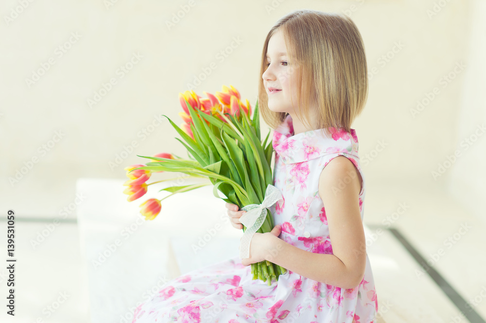 Beautiful toddler girl sitting in the room with fresh tulips, smiling with happy face, waiting for spring and Easter holidays Beautiful toddler girl sitting in the room with fresh tulips, smiling with happy face, waiting for spring and Easter holidays