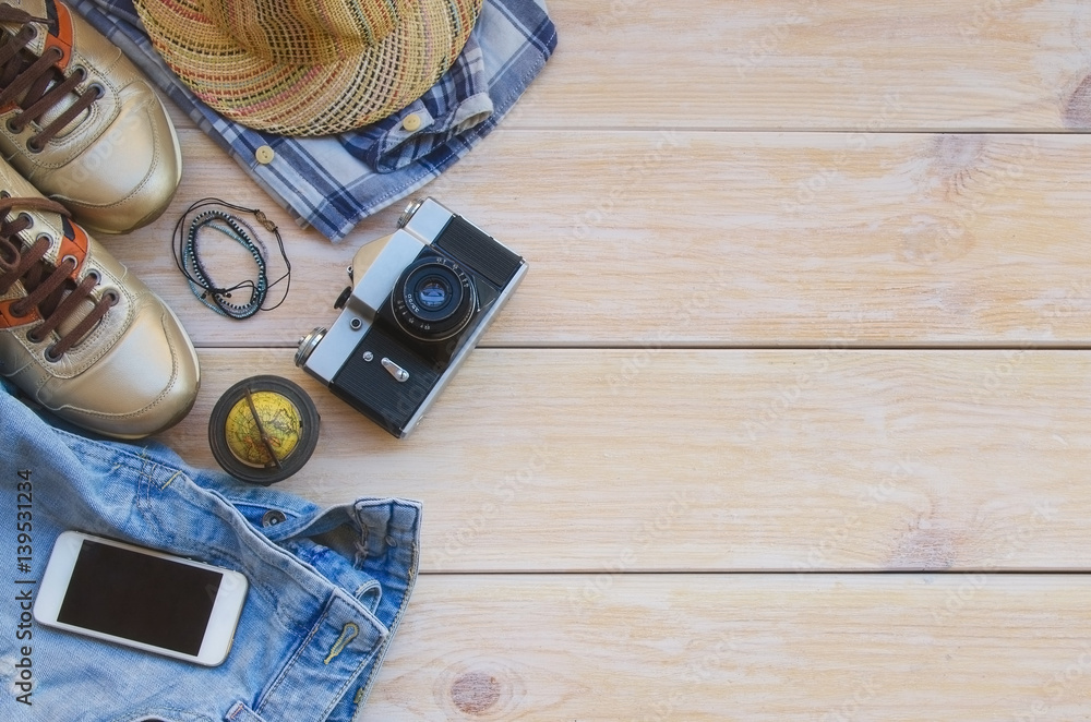 Essential travel items:clothing, smart phone, mini globe and vintage camera  on wooden background. Casual traveler's outfits with blue jeans, hat, check  shirt, gold sneakers and bracelets. Stock Photo