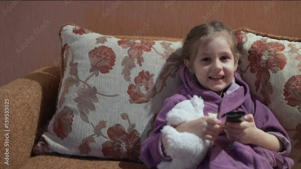 Little girl in violet bathrobe lying on the sofa with teddy bear, watching TV and smiling. Little girl in violet bathrobe lying on the sofa with teddy bear, watching TV and smiling.