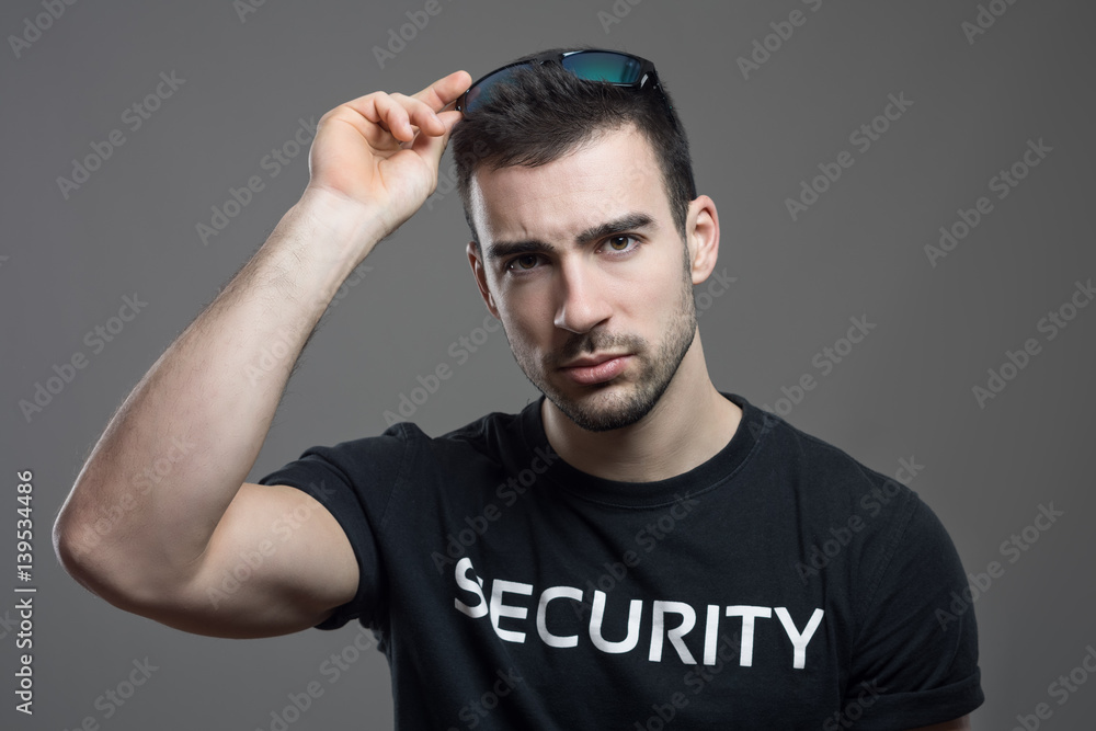 Tough security man taking off sunglasses with intense stare at camera over dark gray studio background. Tough security man taking off sunglasses with intense stare at camera over dark gray studio background.