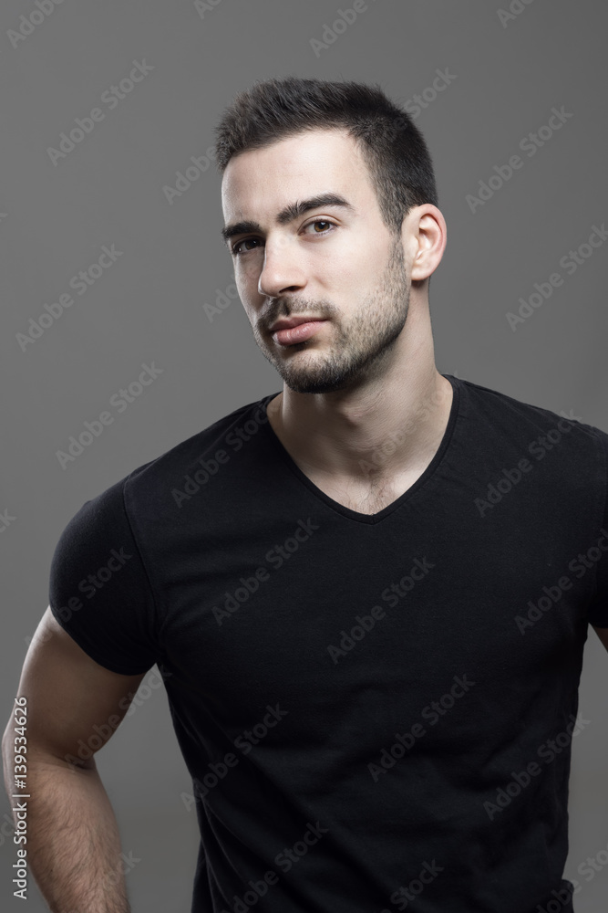 Confident macho athletic man wearing black t-shirt looking at camera suspiciously over dark gray studio background.