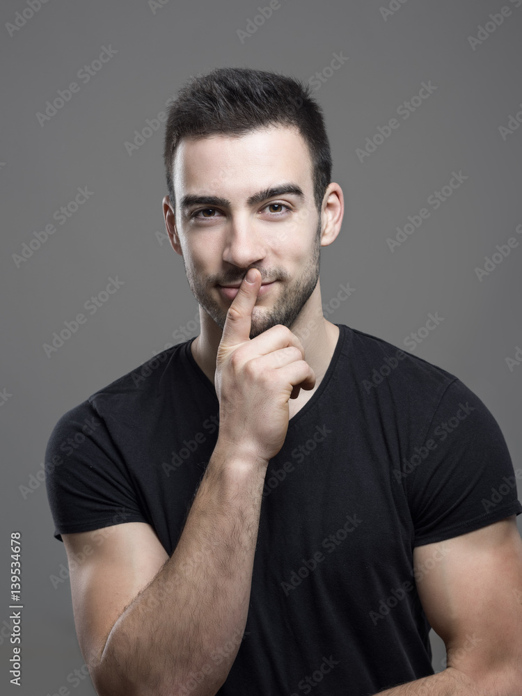 Handsome young fitness male model looking at camera with finger on lips over dark gray studio background. Handsome young fitness male model looking at camera with finger on lips over dark gray studio background.