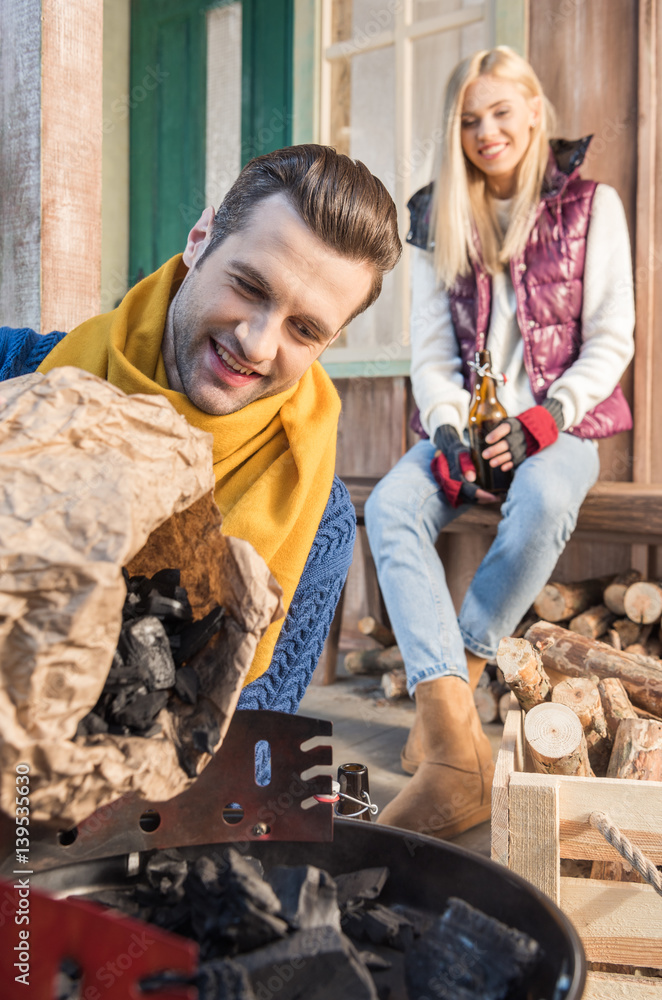 Young woman with beer bottle looking at smiling man filling grill with charcoal Young woman with beer bottle looking at smiling man filling grill with charcoal