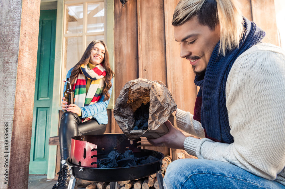 Smiling woman with beer bottle looking at handsome man filling grill with charcoal
