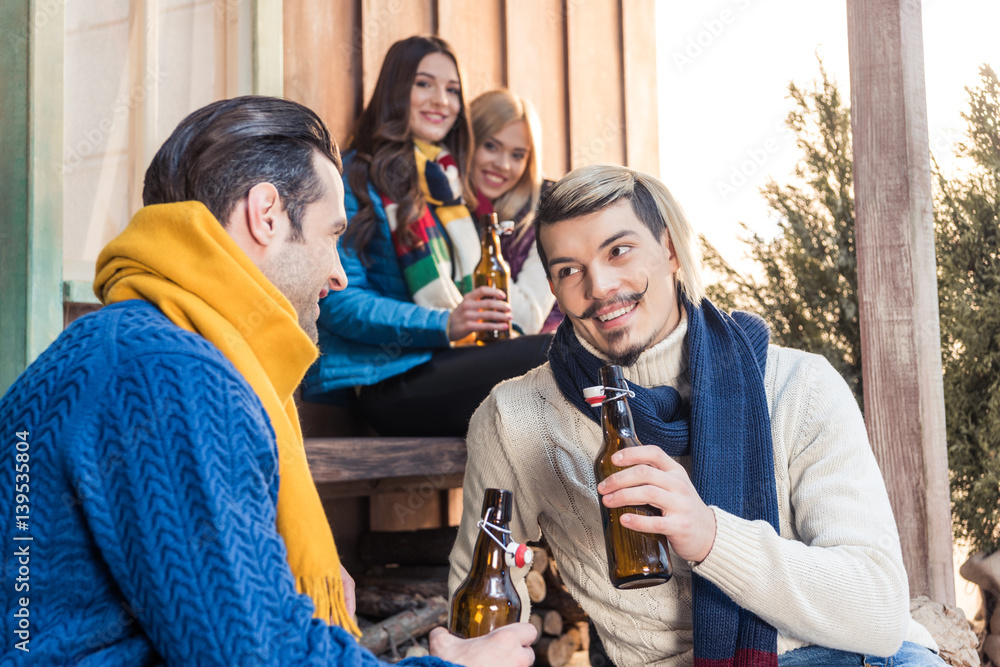 Obraz premium smiling men talking while drinking beer with women looking at them