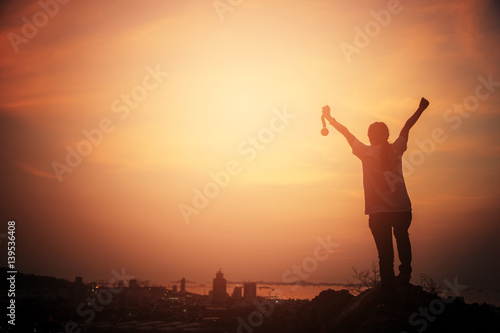 Silhouette woman raising hands and holding gold medal looking the city with sunset sky. victory concept. business success.