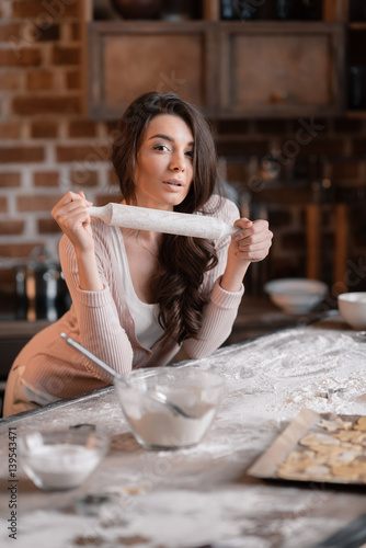  Attractive young woman with rolling pin leaning at kitchen table and looking at camera