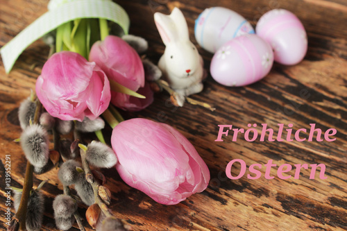 Rosa Tulpen mit Weidekätzchen, Osterhase und Ostereier auf rustikalem Holztisch mit Schriftzug Fröhliche Ostern