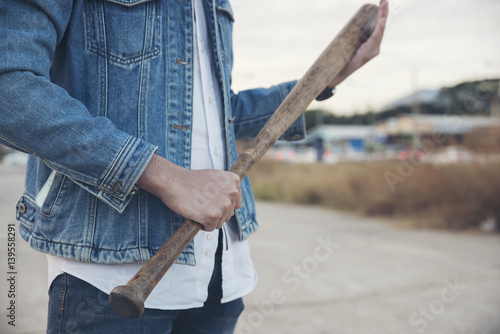 Closed up of smart man wearing blue jeans and jacket standing on the cement patio and holding a baseball bat.