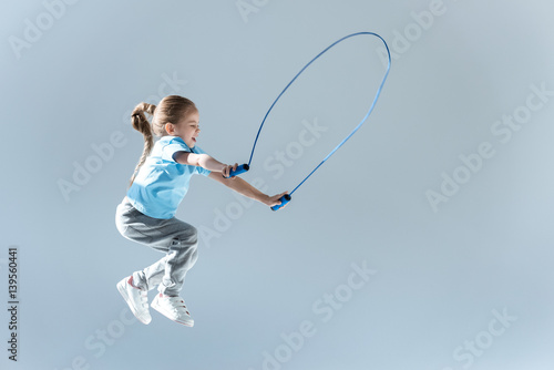 side view of happy girl humping exercising with skipping rope on grey photo