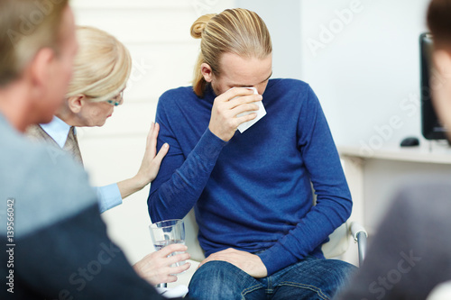 Portrait of young man sitting in circle of psychological support group crying while sharing his trouble with mentor and other participants
