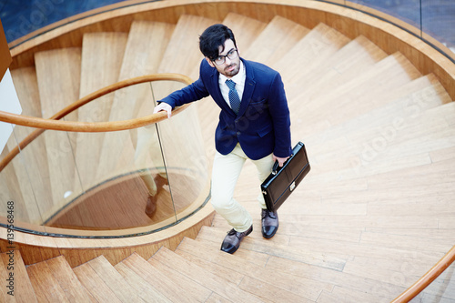 Portrait of modern businessman wearing stylish formal suit going up spiral staircase in designer office building