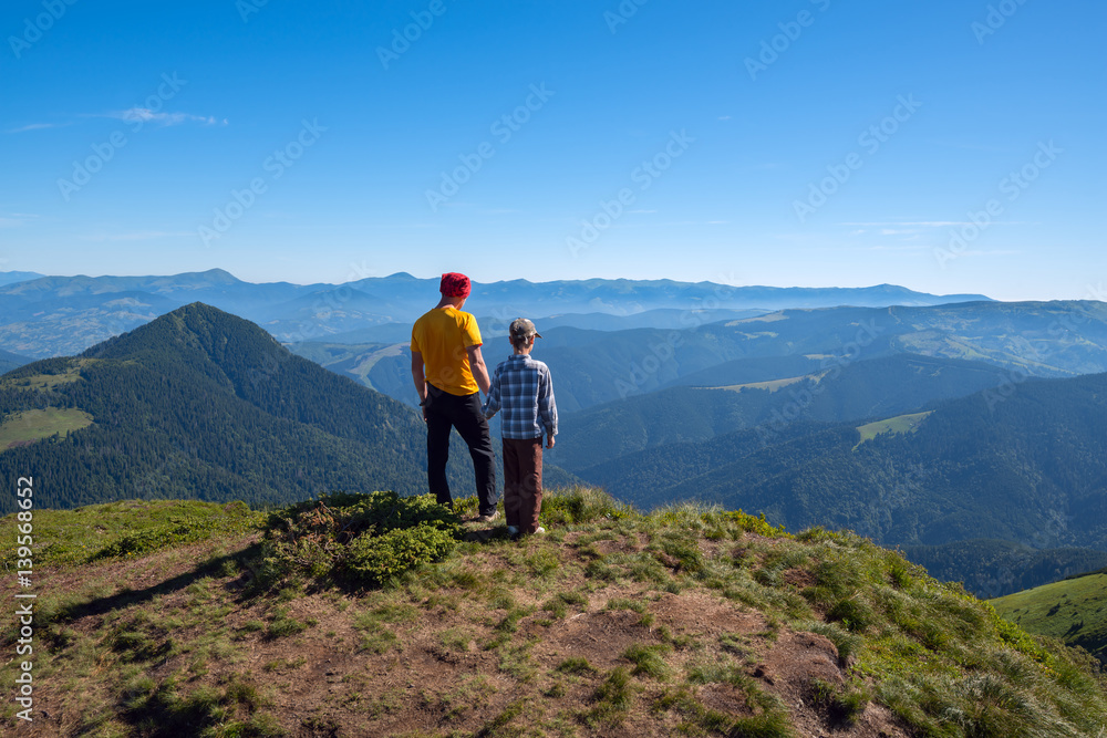 Father and son stand on peak and look into the distance - wonderful adventure in outdoor. Sunny wonderful day in the mountains. Back view.