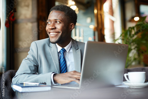 Portrait of handsome African- American businessman wearing formal suit looking away smiling joyfully while busy working with laptop in cafe