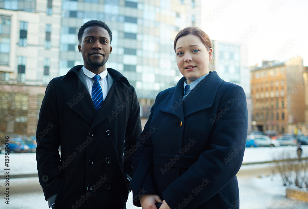 Portrait of two business people, young woman and African-American man, standing outside office building wearing coats and looking at camera Portrait of two business people, young woman and African-American man, standing outside office building wearing coats and looking at camera