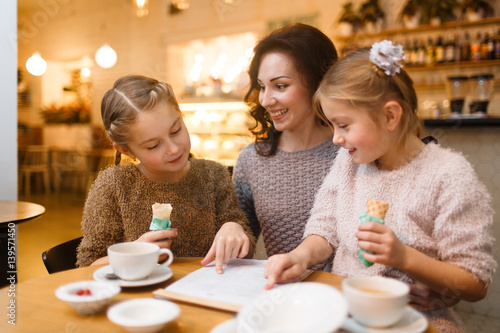Twin girls with ice-cream pointing at document while talking to mother in cafe