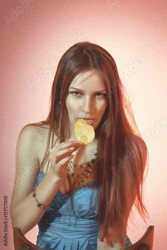 Beautiful colorful toned studio portrait of sensual young woman sitting on the chair and eating potato chip with soulful emotional eyes and long hair photo