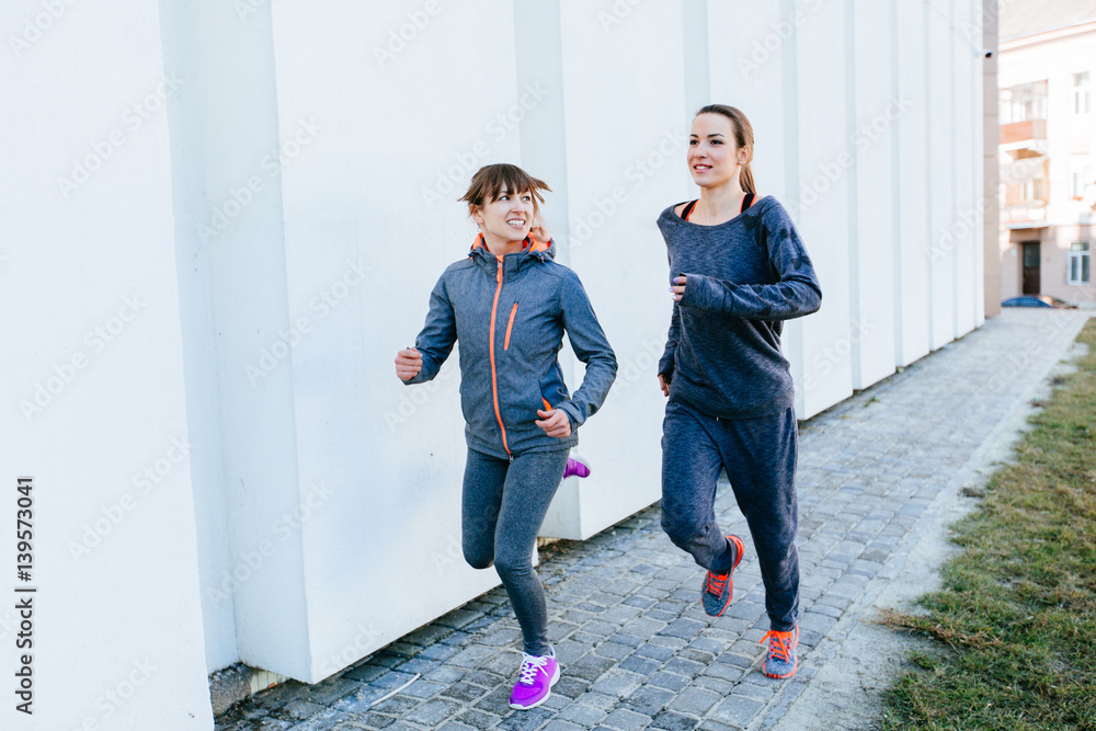 Obraz premium Two sports women in gray sportswear running beside white wall in sunny spring morning