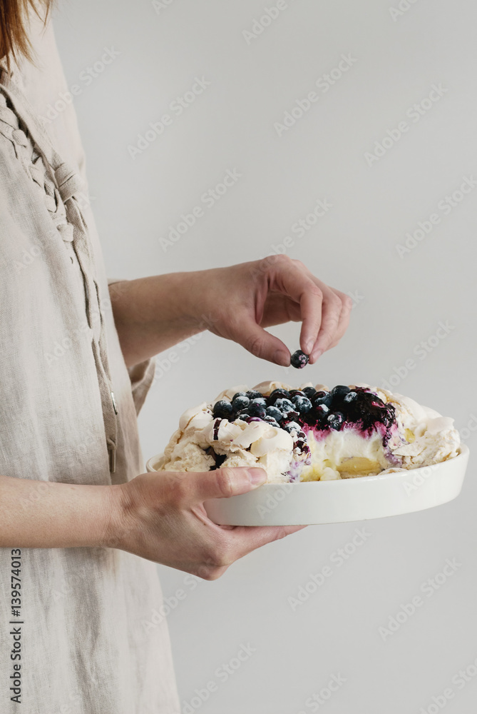 Woman tasting cake. Homemade meringue cake Pavlova with whipped cream, sugar powder, fresh blueberries and blueberry sauce in female hands over gray wall as background, day light.