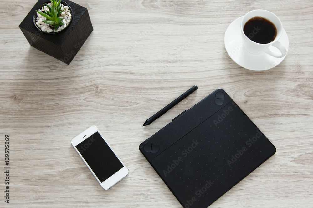 Workplace. On the wooden table there is a graphics tablet, stylus, mobile phone, pot with flowerpots and white cup of coffee. Workplace. On the wooden table there is a graphics tablet, stylus, mobile phone, pot with flowerpots and white cup of coffee.