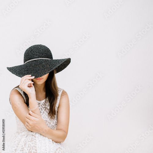 Young pretty woman in white dress with black hat near white wall.
