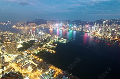 Aerial view of Hong Kong & Kowloon (Tsim Sha Tsui) at night with city skyline of crowded skyscrapers by Victoria Harbour & light trails of ships across seaport~ Cityscape of Hong Kong in blue twilight