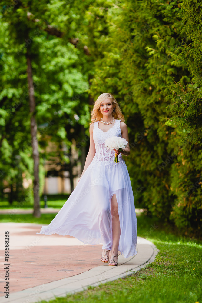 Young beautiful bride in white dress is walking in the summer park with a bridal bouquet 