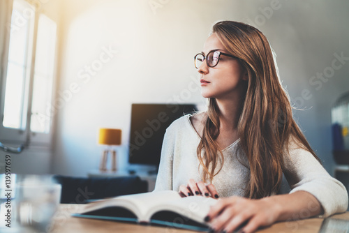 Attractive young girl wearing vintage glasses and reading a book at home interior, hipster girl enjoying sunny day while sitting with a book, pretty woman during a break while working at office